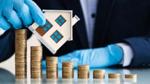 Muž v rukaviciach ukladá mince vedľa modelu domu, symbolizujúc šancu na kúpu nehnuteľnosti za nižšiu cenu.