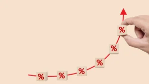 Ruka drží drevený blok s červeným percentuálnym znakom. Značka symbolizuje rastúce úrokové sadzby hypoték, ktoré predstavujú záťaž pre mnohých ľudí.